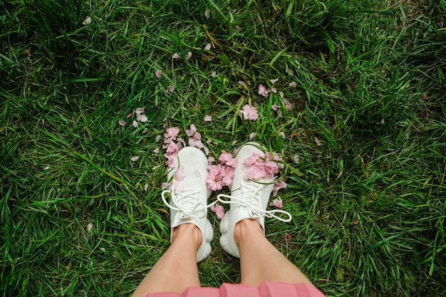 Gambe di donna in scarpe da ginnastica bianche su erba verde con fiori di sakura in fiore rosa copia spazio