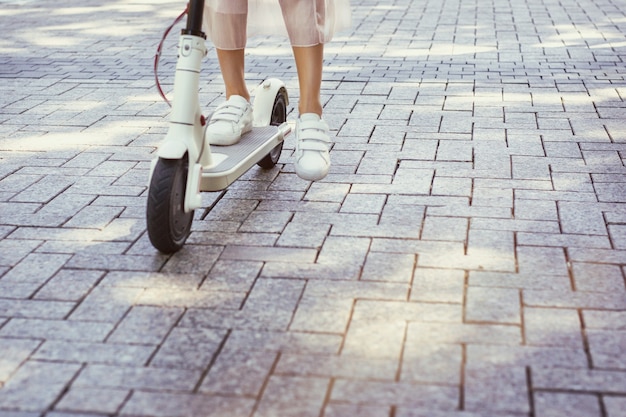 Gambe di donna su scooter elettrico di trasporto ecologico sulla piastrella del marciapiede della città in giornata di sole