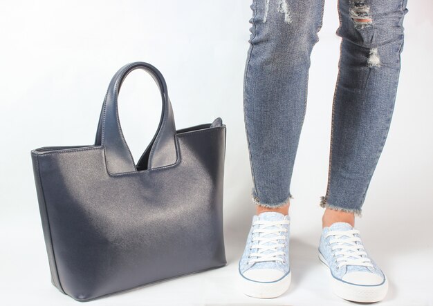 Gambe femminili in jeans e scarpe da tennis che posano con una borsa su bianco