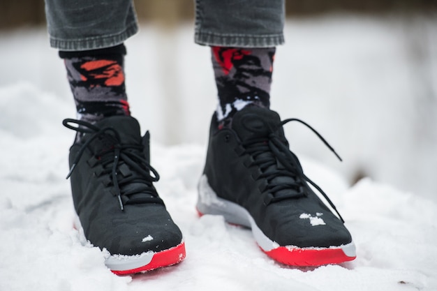 Gambe maschili in scarpe da ginnastica, jeans corti e calzini alla moda in piedi sulla neve.