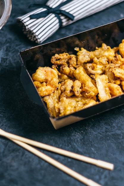 Gamberi, calamari e polpi cotti in pastella in una scatola di carta nera su fondo scuro.