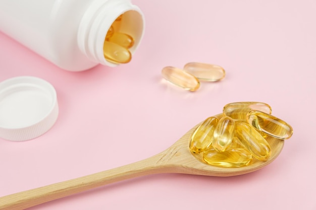 Gelatina di pesce Softgels Omega-3 in cucchiaio di legno e barattolo di plastica bianco aperto con capsule su sfondo rosa