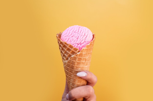 Gelato in tazza di cialda in mano femminile su sfondo giallo. Gelato alla fragola rosa in un cono di cialda