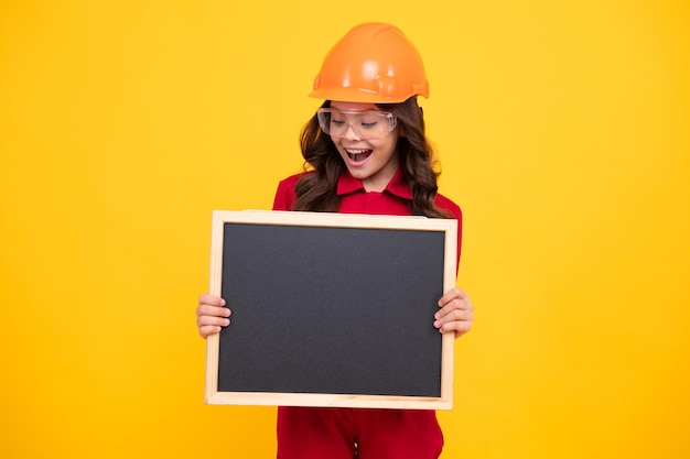 Generatore di ragazza bambino nel casco elmetto Lavoratore ragazza adolescente tenere lavagna isolata su sfondo giallo Concetto di ristrutturazione per bambini Copia spazio mock up