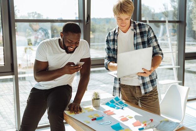 Generazione moderna. Allegro afro american uomo utilizzando il suo smart phone e seduto in ufficio mentre il suo allegro collega utilizzando il computer portatile