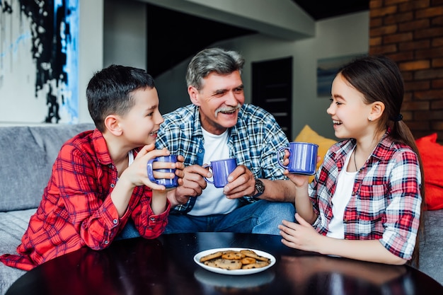 Generazioni felici. Uomo anziano sorridente allegro che beve tè con un biscotto mentre si gode il tempo con i suoi nipoti.