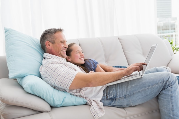 Generi con la figlia che si rilassa sullo strato facendo uso del computer portatile a casa nel salone