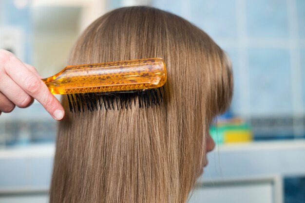 Generi la mano con la spazzola che pettina i capelli biondi lunghi della ragazza sveglia del bambino dopo il bagno su fondo interno vago.