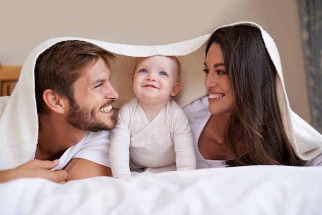 Genitori di famiglia felici e bambino con coperta sul letto per la cura dell'amore e del tempo di qualità insieme Madre padre e bambino appena nato giocoso che si rilassano in camera da letto con biancheria da letto forte sorriso e legame a casa