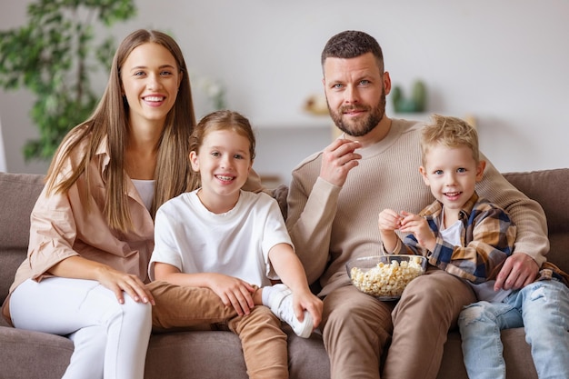 Genitori e bambini felici della famiglia che mangiano popcorn e ridono seduti sul divano e guardano film divertenti a casa