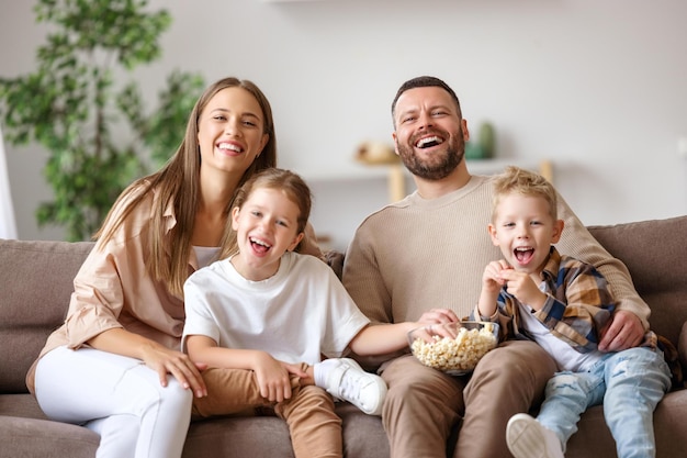 Genitori e bambini felicissimi che mangiano popcorn e ridono seduti sul divano e guardano film divertenti a casa