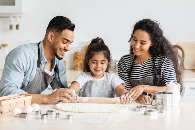 Genitori e bambini sorridenti della famiglia mediorientale che preparano biscotti insieme a casa, rotolano la pasta e conversano, primo piano. Felice padre arabo, madre e figlia che cuociono in cucina