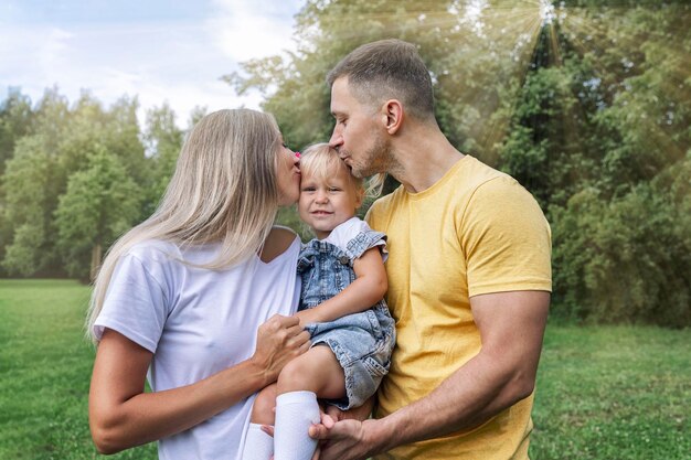 Genitori felici con la loro piccola figlia nel parco in una soleggiata giornata estiva Ridendo giovane donna con un uomo e un bambino in jeans e magliette abbraccio e bacio Amore e tenerezza