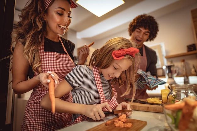 Genitori felici e la loro figlia che cucinano insieme in cucina mentre la bambina cerca di tagliare la carota.