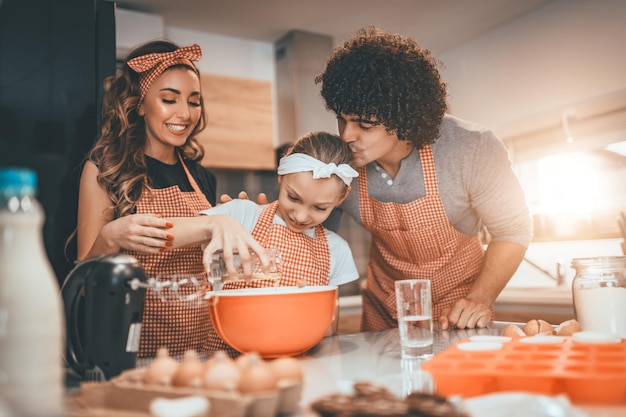 Genitori felici e la loro figlia stanno preparando i biscotti insieme in cucina. La bambina aiuta i suoi genitori a versare l'olio nel piatto con l'impasto.