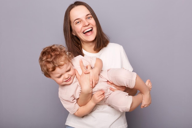 Genitorialità felice Ridendo bella donna che si diverte con il suo bambino neonato che gioca godendosi il tempo insieme in piedi isolato su sfondo grigio