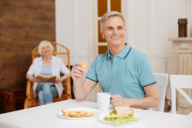 Gentiluomo bello entusiasta positivo seduto al tavolo e godersi il suo pasto mattutino guardando fuori dalla finestra