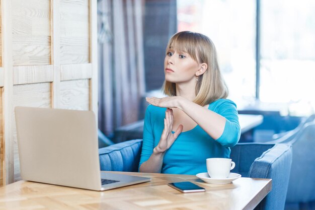 Gesto del tempo! Ritratto di vista laterale di avvertimento attraente giovane freelancer con i capelli biondi in camicetta blu sono seduti al bar e fare videochiamate sul computer portatile con le braccia alzate e mostrando la pausa