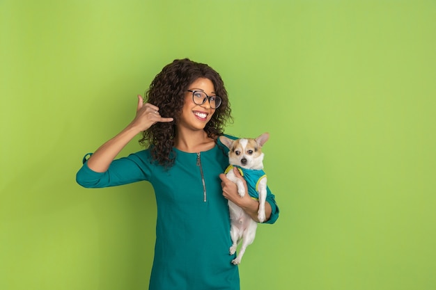 Gesto di chiamare il telefono. Ritratto di giovane donna afro-americana isolato su sfondo verde studio. Bellissimo modello con cagnolino. Concetto di emozioni umane, espressione facciale, vendite, pubblicità.