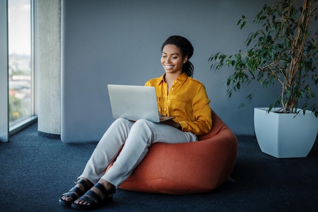 Gestore femminile nero positivo che lavora al computer portatile in ufficio seduto sullo spazio libero della sedia del pouf beanbag