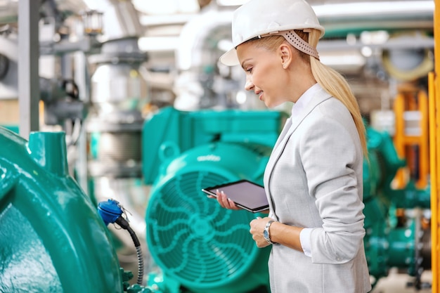 Gestore femminile sorridente riuscito laborioso in vestito che sta nella centrale termica con il ridurre in pani nelle mani e nel controllo sulla turbina.