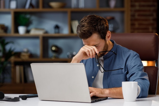 Gestore maschio bello dell'uomo d'affari che lavora stancamente dietro un computer portatile in un ufficio domestico alla moda perdita di v