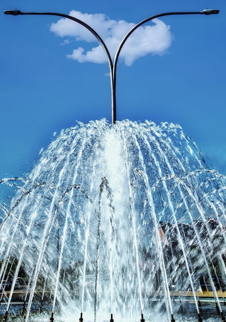Getti d'acqua da una fontana con un lampione e una piccola nuvola dietro.