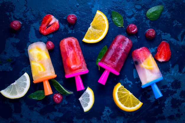 Ghiacciolo di frutta brillante fatto in casa con aroma di fragola, ciliegia, limone, arancia, limone e menta e frutta fresca per gelato su una superficie blu scuro. Vista piana, vista dall'alto