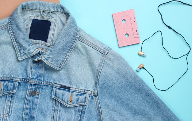 Giacca di jeans, audiocassetta, auricolari aspiranti su superficie blu pastello. Media retrò, amante della musica, anni '80. Vista dall'alto, piatto