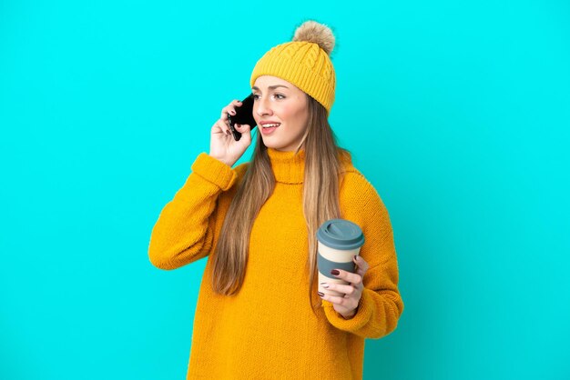 Giacca invernale da portare della giovane donna caucasica isolata su fondo blu che tiene il caffè da portare via e un cellulare