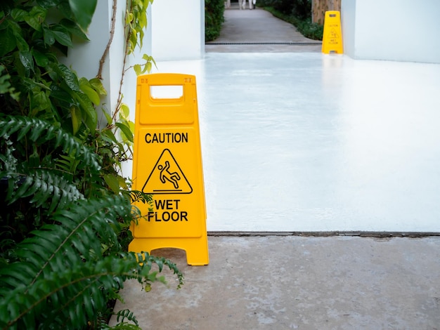 Giallo attenzione segno pavimento bagnato scivoloso con persona scivolosa icona di avvertimento su sfondo pavimento di cemento bianco bagnato di fronte all'edificio vicino giardino di foglie verdi dopo la pioggia con lo spazio della copia