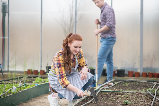 Giardinaggio. Donna sorridente in grembiule accovacciata vicino al letto del giardino uomo con rastrello in piedi dietro