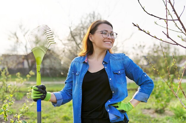 Giardinaggio primaverile, ritratto di donna matura sorridente giardiniere con rastrello. Sfondo di alberi e cespugli, pulizia stagionale dell'erba e delle foglie dell'anno scorso in giardino, cortile