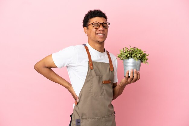 Giardiniere africano che tiene una pianta isolata su sfondo rosa che soffre di mal di schiena per aver fatto uno sforzo