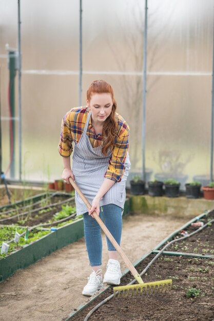 Giardiniere. Coinvolta giovane donna adulta con rastrello che lavora vicino al letto del giardino nella sua serra