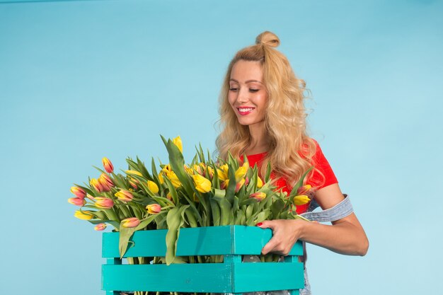 Giardiniere della bella donna bionda che tiene scatola con i tulipani sulla parete blu