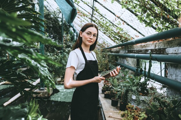 Giardiniere della giovane donna con tavoletta digitale che lavora in serra