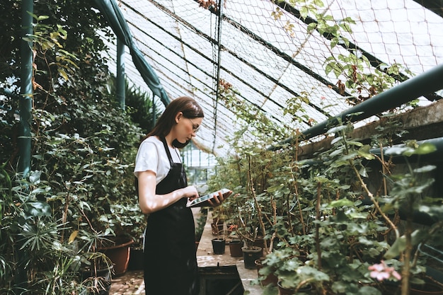 Giardiniere della giovane donna con tavoletta digitale che lavora in serra