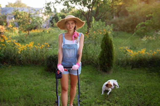 Giardiniere delle donne. Una ragazza sexy con un cappello falcia l'erba vicino alla casa con un tosaerba. Progettazione del paesaggio e cura del prato