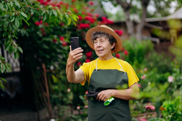 Giardiniere donna senior in un cappello che lavora nel suo cortile e parla al telefono Il concetto di giardinaggio che cresce e si prende cura di fiori e piante