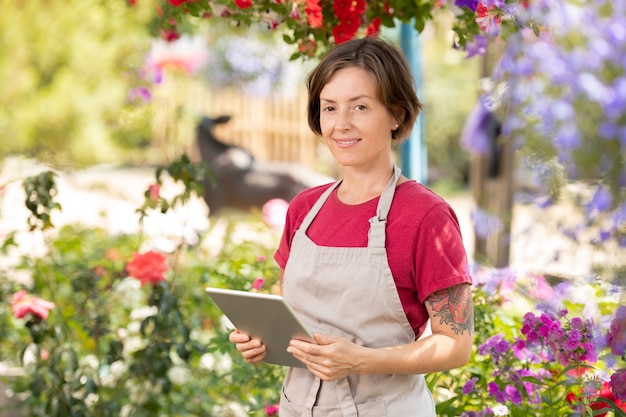 Giardiniere femminile felice con touchpad in piedi in serra e lavorando sul listino prezzi online per le piante sul sito web