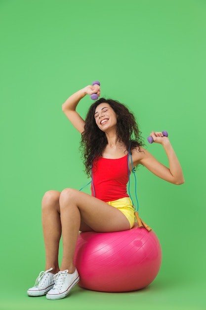 Ginnastica donna che indossa abiti estivi sollevamento manubri mentre è seduto sulla palla fitness durante l'aerobica contro il muro verde