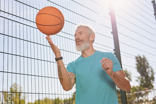 Giocare a basket all'aperto felice uomo anziano in abbigliamento sportivo che fa girare una palla da basket sul suo