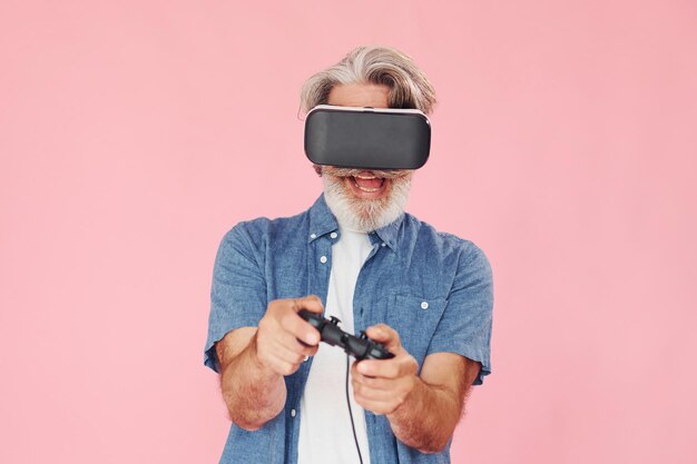 Giocare con occhiali per realtà virtuale e joystick L'uomo anziano moderno ed elegante con i capelli grigi e la barba è al chiuso