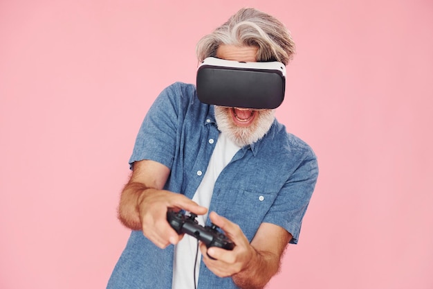 Giocare con occhiali per realtà virtuale e joystick L'uomo anziano moderno ed elegante con i capelli grigi e la barba è al chiuso