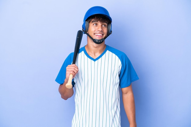 Giocatore di baseball caucasico con casco e mazza isolato su sfondo blu pensando a un'idea mentre guarda in alto