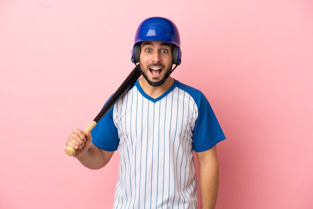 Giocatore di baseball con casco e mazza isolato su sfondo rosa con espressione facciale a sorpresa