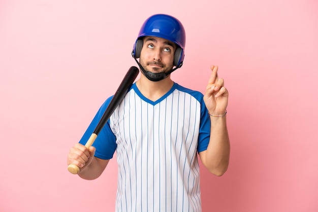 Giocatore di baseball con casco e mazza isolato su sfondo rosa con le dita incrociate e augurando il meglio