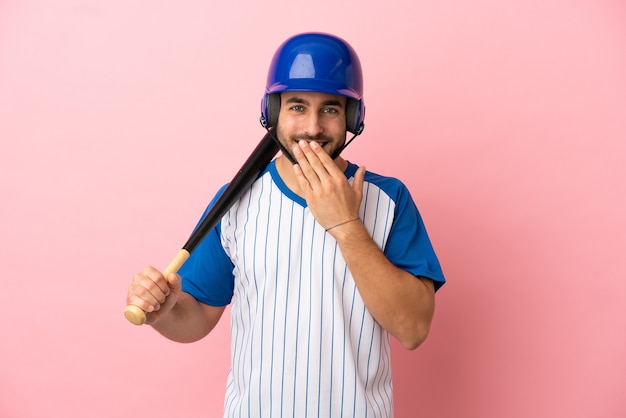 Giocatore di baseball con casco e mazza isolato su sfondo rosa felice e sorridente che copre la bocca con la mano