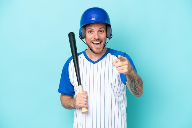 Giocatore di baseball con casco e pipistrello isolato su sfondo blu sorpreso e puntato davanti